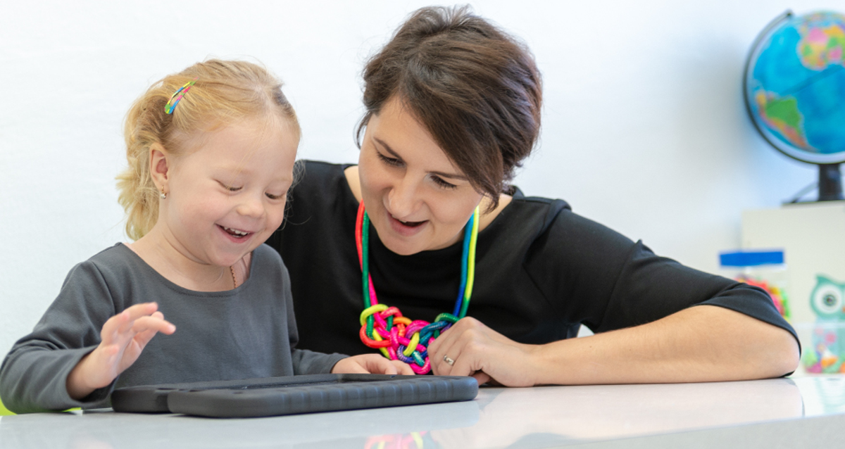An Image of a toddler girl with disability alongside her therapist, on a electronic device