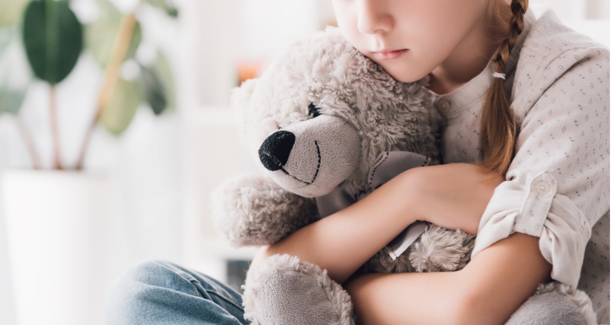 A young girl with a plait in her hair and a thoughtful look on her face is hugging a teddy bear. The teddy is smiling.