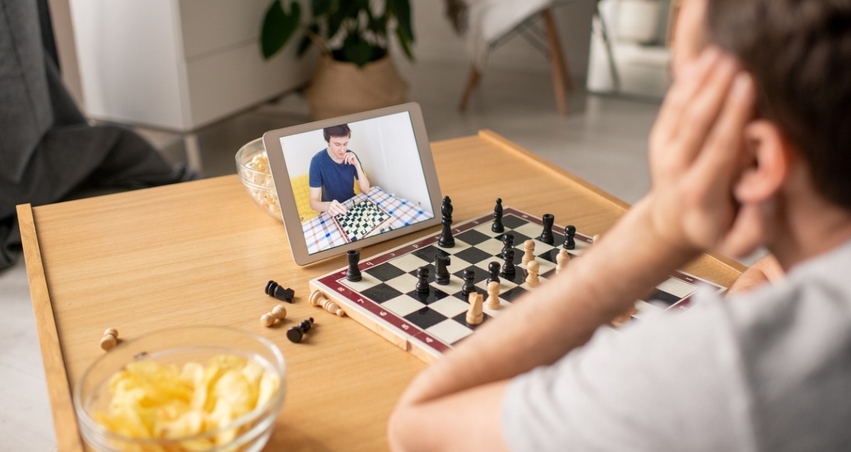 A boy sits with a chess board and ipad in front of him. A bowl of chips is to the side. On the ipad is a friend he is playing against, who also has a chessboard that we can see in the picture.