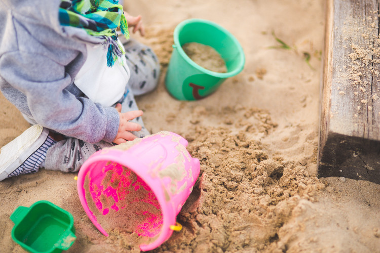Sand play and sand pits. A little boy is in a sandpit, with buckets beside him.