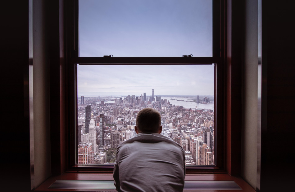 Man in grey shirt looking out window at city buildings