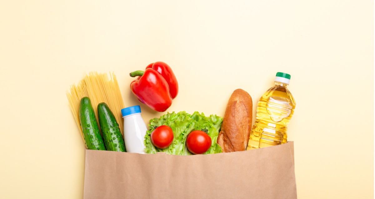 An image of a paper bag with cucumber, pasta, milk, capsicum, lettuce, tomato, bread and oil peeking out of the top. A concept photo of a food hamper.