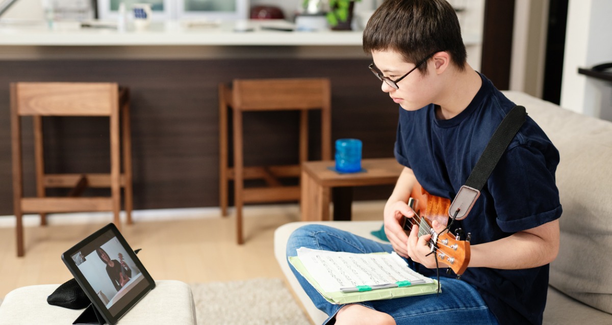 Teenage boy with down syndrome taking music lessons over the internet. He sits with music in his lap, is holding his instrument and is watching his teacher on a device