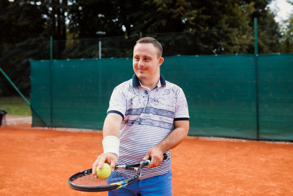 An Image of a young man with disability, holding a tennis racquet and ball.