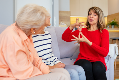 An image of three women siting and communicating, one is using sign language and the others are looking on.,
