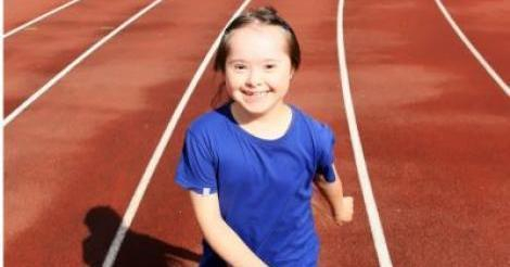 A photo of a young girl with down syndrome smiling at the camera. She is standing on an athletics track.